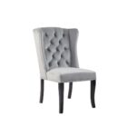 Grey velvet dining chair with a button-tufted wingback, padded seat, stud trim detailing, and curved black wood legs.